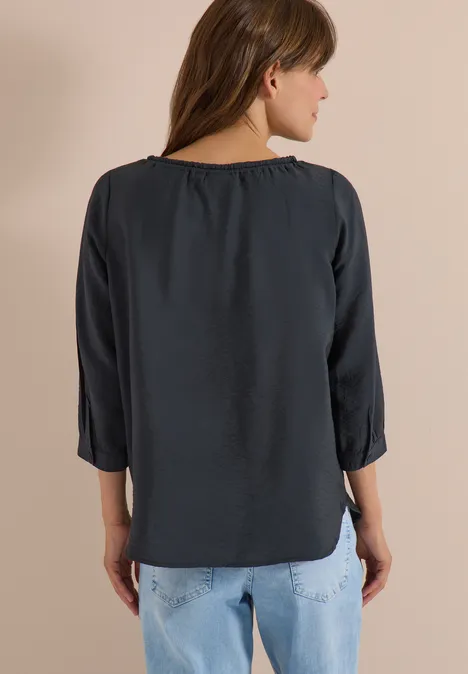 Frau steht von hinten, trägt dunkelgraue 3/4-Ärmel-Bluse und helle Jeans.