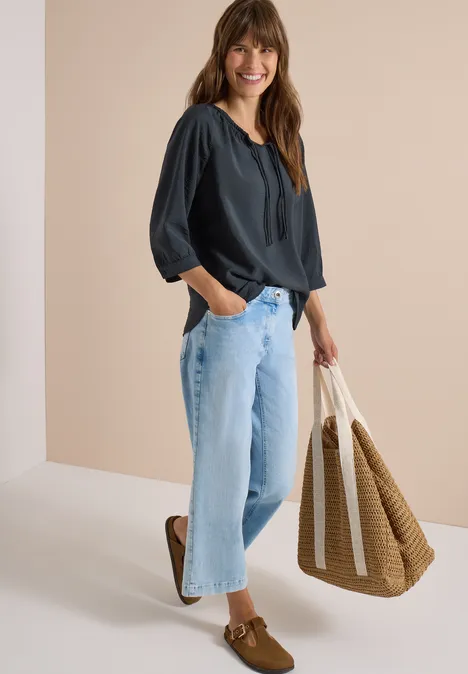 Frau in dunkelblauer Bluse, hellen Jeans und Loafers trägt geflochtene Tasche und lächelt vor beigem Hintergrund.
