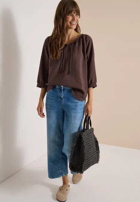 Frau in brauner Bluse, weiten Jeans und Sneakers trägt schwarze geflochtene Tasche und lächelt.
