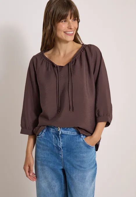 Lächelnde Frau in brauner Bluse und blauen Jeans, eine Hand in der Hosentasche.