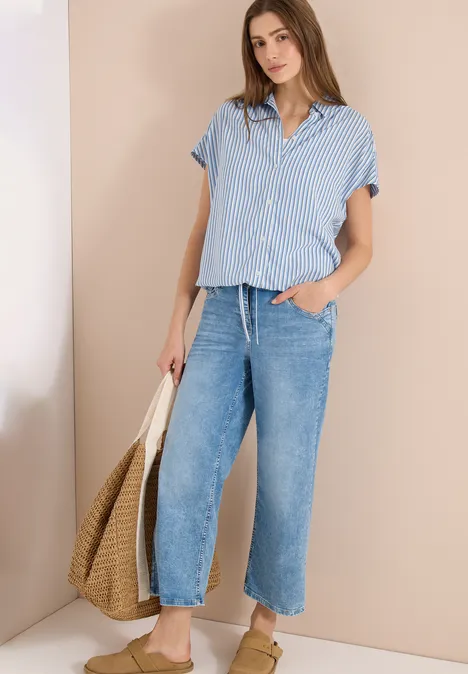 Frau in blau-weiß gestreiftem Kurzarmhemd, weiten Jeans und Strohtasche vor beigem Hintergrund.