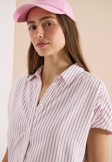 Eine Person trägt eine rosa Kappe und ein weiß-rosa gestreiftes Hemd vor beigem Hintergrund.