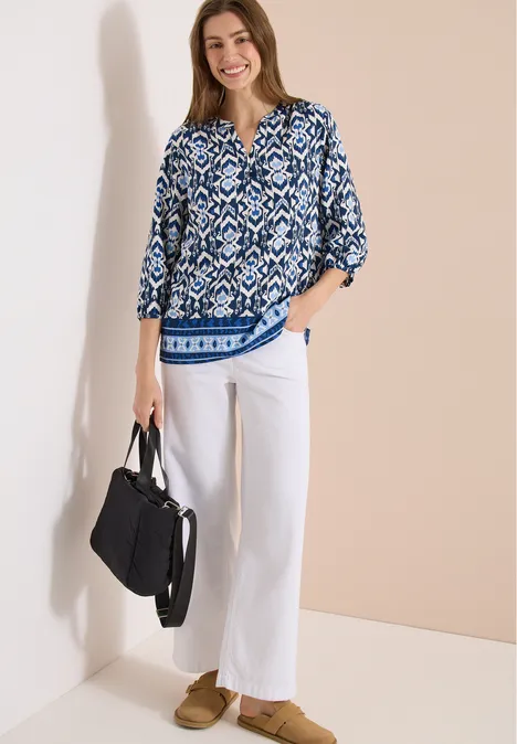 Frau trägt blau-weiß gemusterte Bluse, weiße Hose, beiger Loafer und schwarze Handtasche vor hellem Hintergrund.