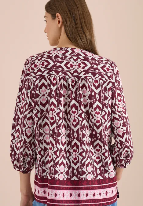 Rückansicht einer Frau in locker geschnittener, rot-weiß gemusterter Bluse mit 3/4-Ärmeln vor beigem Hintergrund.