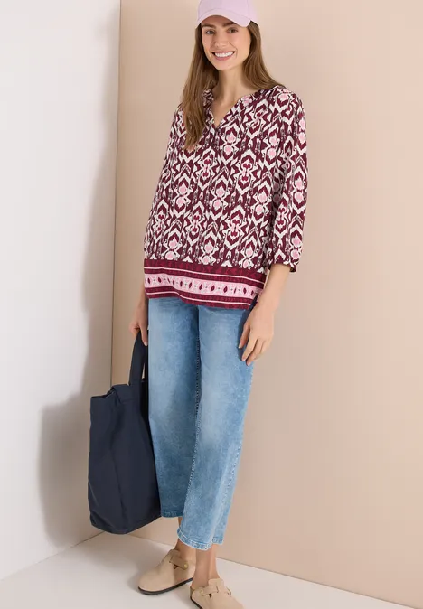 Frau trägt bunte V-Ausschnitt-Bluse, weite Jeans, bequeme Schuhe und hält eine blaue Stofftasche.