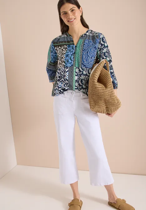 Frau trägt blaue gemusterte Bluse, weiße Schlagjeans, Strohtasche und braune Sandalen.