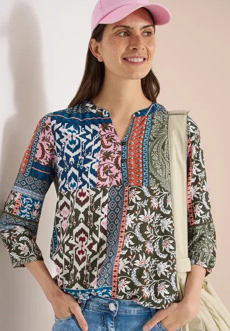 Frau trägt bunte, gemusterte Bluse, Jeans, pinke Baseball-Cap und Schultertasche.