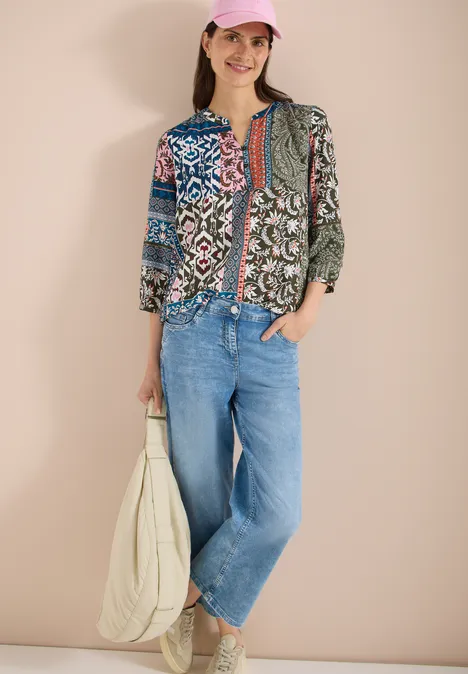 Frau vor beigem Hintergrund trägt bunte Patchwork-Bluse, weite Jeans und weiße Tasche.