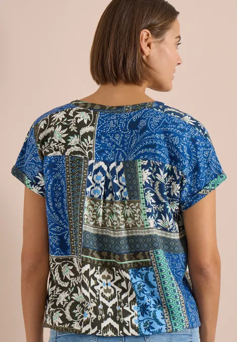 Rückansicht einer Frau, die ein kurzärmeliges Oberteil mit blau-grünem Patchwork aus floralen und geometrischen Mustern trägt.