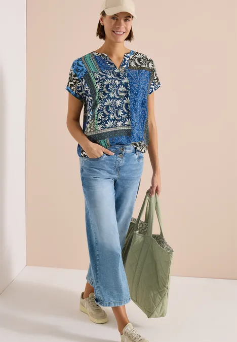 Eine Frau trägt eine gemusterte blaue Bluse, helle Cropped-Jeans, beigen Sneaker und hält eine große grüne Tragetasche.