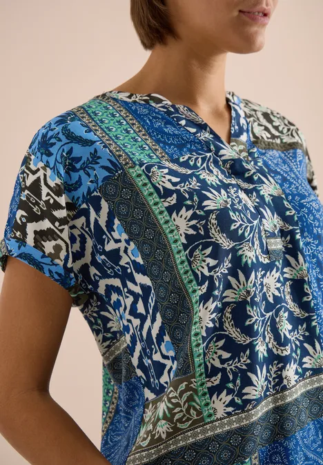 Frau in kurzärmliger, blau-grün gemusterter Patchwork-Bluse vor neutralem Hintergrund.