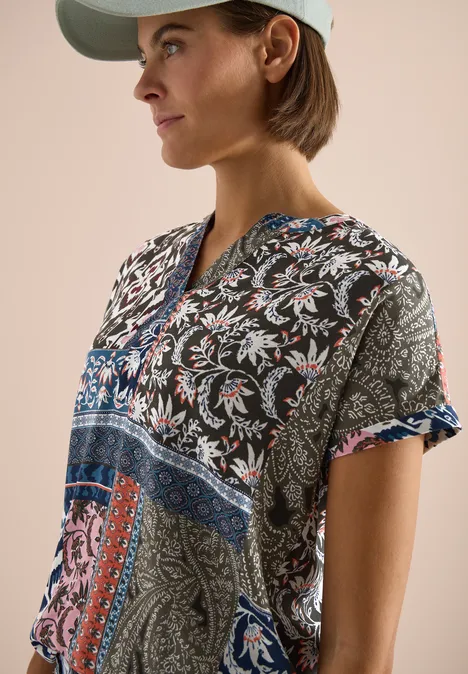 Iemand met pet draagt een kleurrijke paisley-tuniek met V-hals en korte mouwen voor een beige achtergrond.