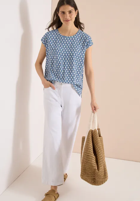 Frau trägt blau gemustertes Shirt, weiße Schlaghose und große Strohtasche.