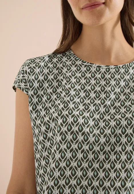 Een vrouw draagt een groen topje met korte mouwen en een wit-groen ruitpatroon tegen een beige achtergrond.