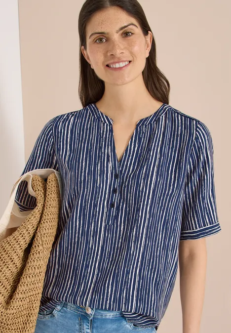 Frau in blau-weiß gestreiftem Hemd und weißer Hose hält eine Korbtasche vor hellem Hintergrund.