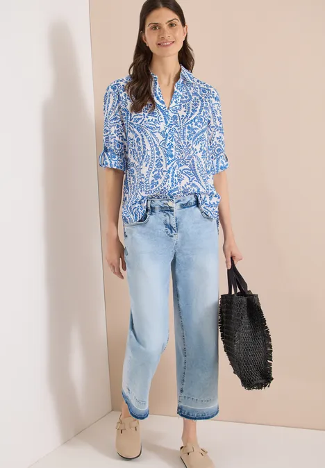 Frau lehnt an Wand, trägt blau gemusterte Bluse, weite Jeans, bequeme Schuhe und schwarzen Flechtkorb.