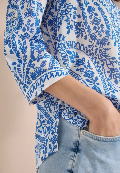 Frau trägt weiße Bluse mit blauem Paisley-Muster, Hand in der Tasche ihrer hellblauen Jeans.