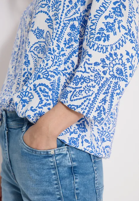 Ausschnitt einer Person in weiß-blau gemusterter Bluse, Hand in Jeanshose gesteckt.