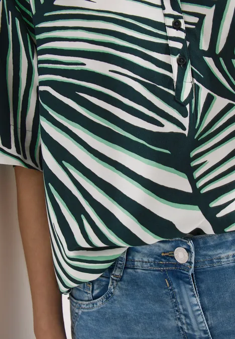 Person trägt weiß-grünes, palmblattgemustertes Oberteil und blaue Jeans.