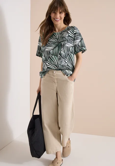 Frau trägt grün-weißes Palmendruck-Oberteil, helle weite Hose, Beuteltasche, bequeme Schuhe vor beiger Wand.