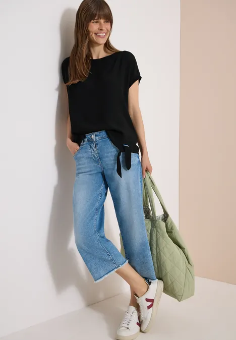 Frau in schwarzem T-Shirt, weitem Jeans-Culotte, weiß-roten Sneakern und grünem Stoffbeutel vor hellem Hintergrund.