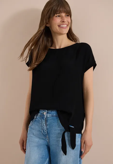 Eine Frau trägt eine schwarze Bluse mit seitlichem Knoten und blaue Jeans.
