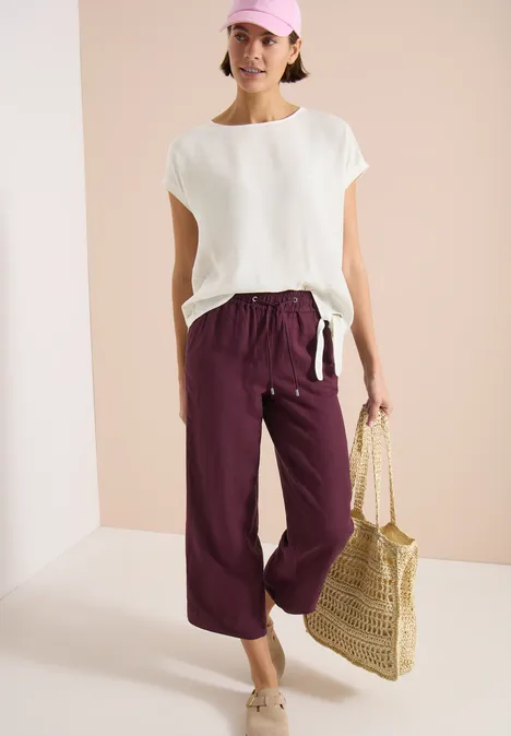 Blouse met ronde hals en knoopdetail vanilla white Vrouw in een wit topje, wijde bordeauxrode broek met strooien tas.