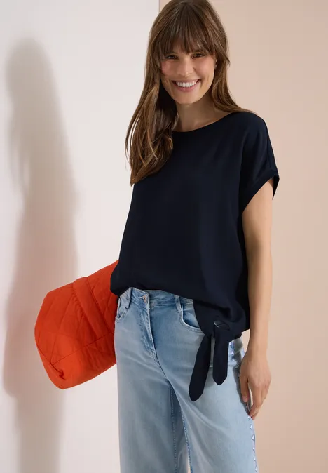 Frau in dunkelblauem, seitlich geknotetem T-Shirt, hellen Jeans, roter Tasche lässig unter dem Arm.
