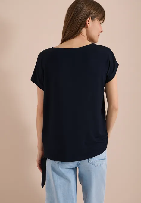 Frau von hinten in dunkelblauem T-Shirt und hellblauen Jeans vor neutralem Hintergrund.