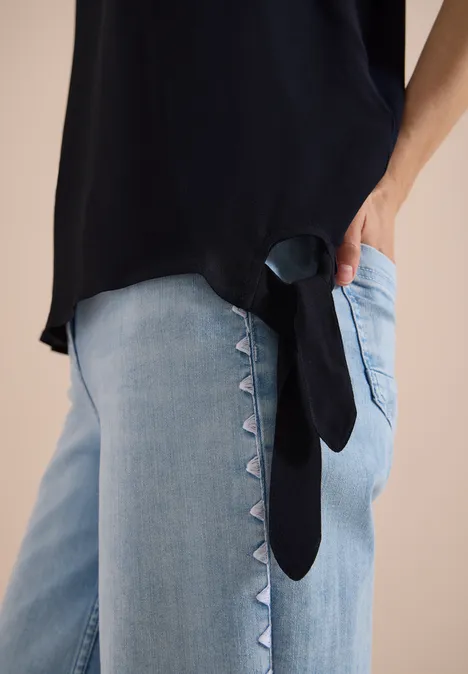 Schwarzes Top und helle Jeans mit seitlichem Dreieckbesatz, Hand in der Tasche.