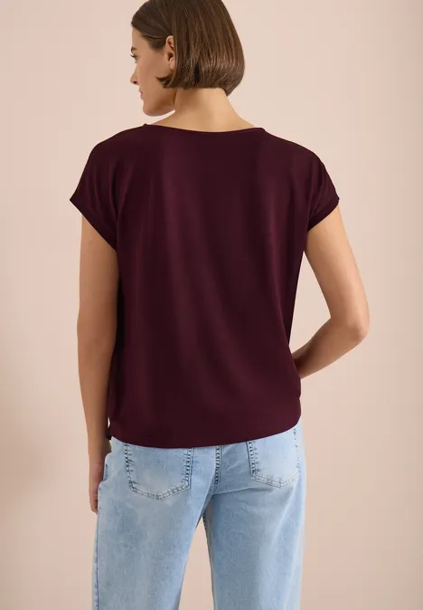 Bluse mit Rundhals und Knotendetail sweet grape red Rückansicht einer Frau in bordeauxrotem Kurzarmshirt und hellblauen Jeans vor neutralem Hintergrund.