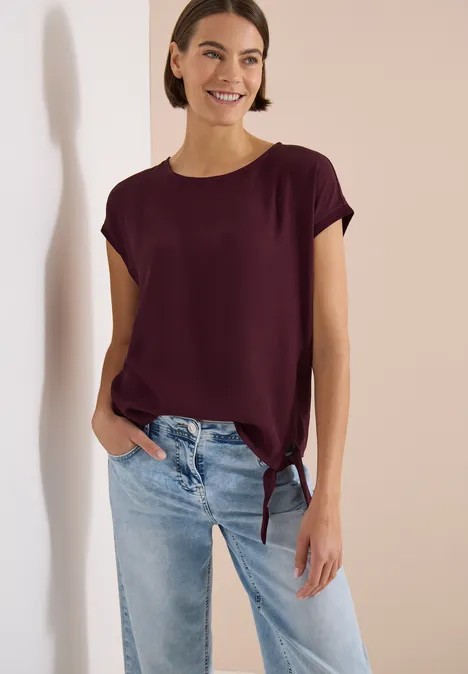 Bluse mit Rundhals und Knotendetail sweet grape red Eine Frau in dunkelrotem T-Shirt mit seitlicher Schleife und hellen Jeans.