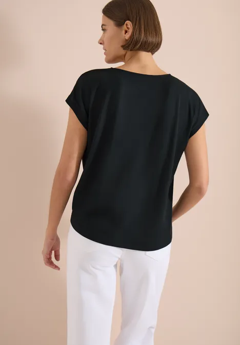 Eine Frau von hinten in schwarzem T-Shirt und weißen Hosen vor beigem Hintergrund.