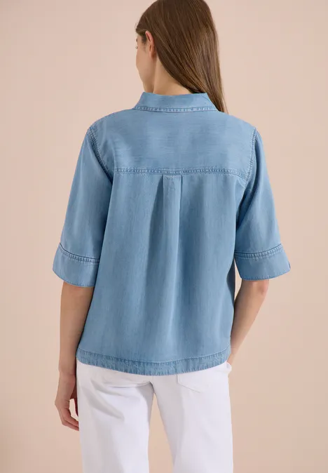 Rückansicht einer Frau in hellblauer Jeansbluse und weißer Hose.