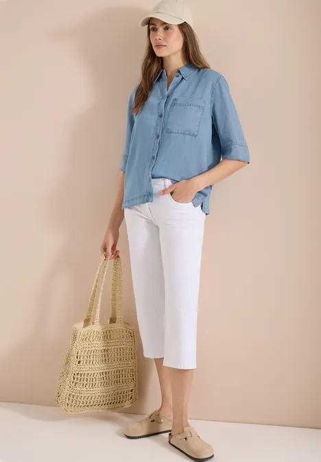 Frau trägt hellblaues Hemd, weiße Culottes, beigefarbene Ballerinas und hält eine geflochtene Strohtasche.