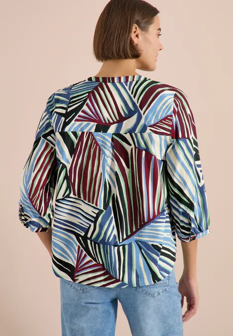 Vrouw van achteren in losvallende blouse met geometrische strepen in blauw, groen, rood en wit.
