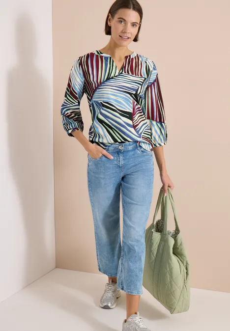 Vrouw met kleurrijk gestreepte blouse, blauwe jeans en groene tas voor een lichte muur.