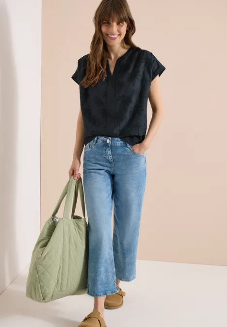 Frau trägt schwarzen Top, weite blaue Jeans, hält großen olivgrünen Shopper, beige Schuhe.
