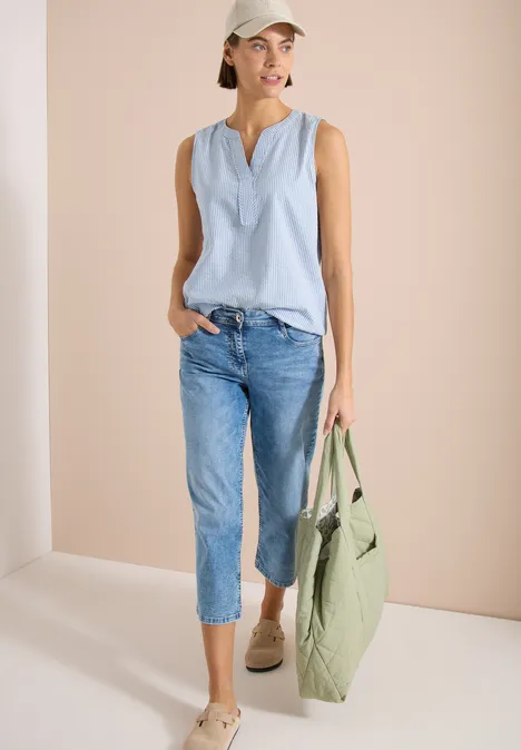 Femme en haut sans manches bleu clair, jean et espadrilles tenant un grand sac vert sur fond sobre.