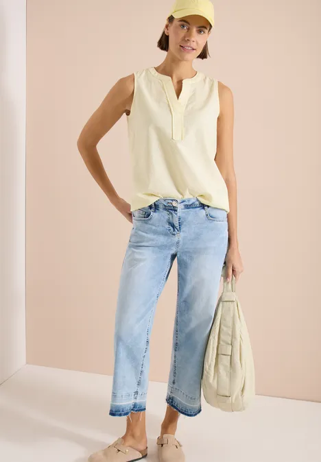 Frau in ärmellosem cremefarbenem Top, hellblauen Jeans und passender Tasche vor neutralem Hintergrund.