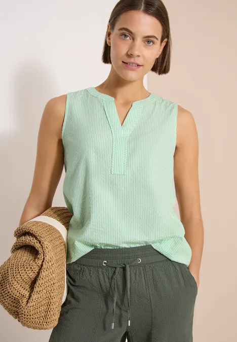 Femme en haut sans manches vert menthe, pantalon foncé, tenant un chapeau de paille.