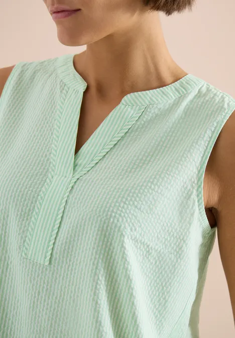 Femme portant un top sans manches vert pâle, col montant et encolure en V, tissu léger gaufré.