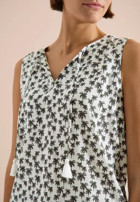 Vrouw draagt een mouwloze witte top met groen palmboomdessin en kwastjes bij de V-hals.