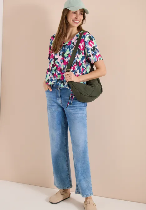 Vrouw in bloemenblouse, rechte spijkerbroek en groene schoudertas tegen beige achtergrond.