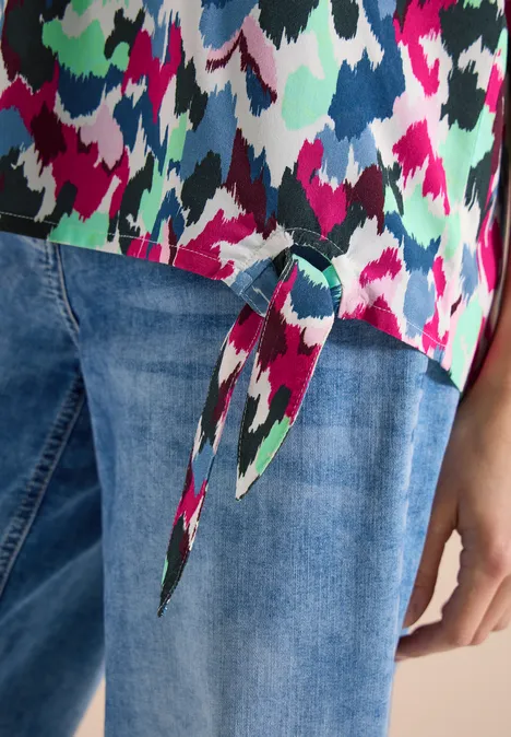 Kleurrijke top met abstract patroon en knoop in de zoom, gecombineerd met een spijkerbroek.