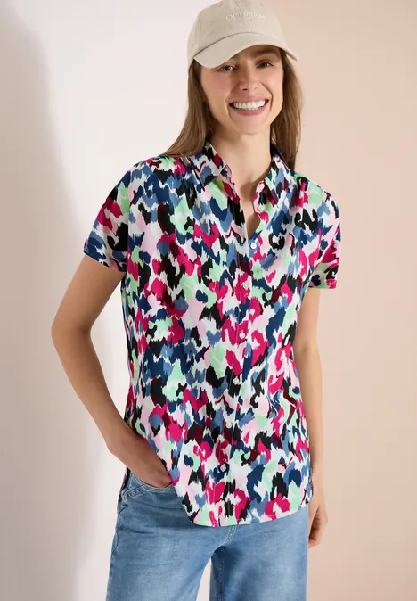 Frau trägt ein kurzärmliges, buntes Camouflage-Hemd und Jeans, Hände in den Taschen, vor hellem Hintergrund.