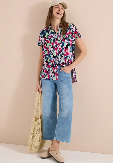 Eine Frau trägt eine bunte Kurzarmbluse, vorn geknotet, weite Jeans, Basttasche und Slipper.