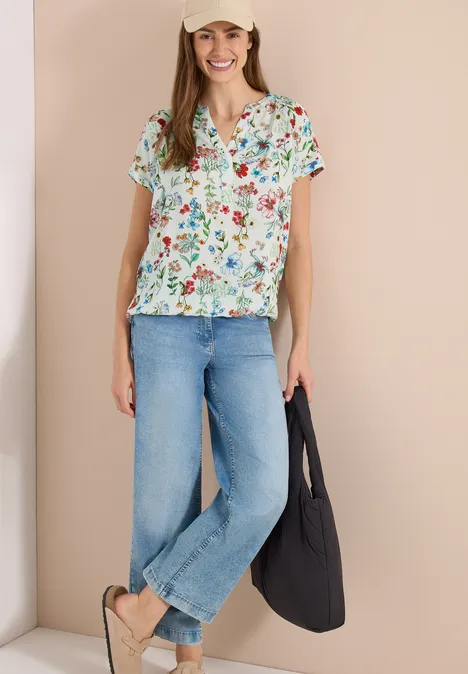 Frau in bunter Bluse mit Blumenmuster, weiter blauer Jeans, Sandalen und schwarzer Tasche vor hellem Hintergrund.