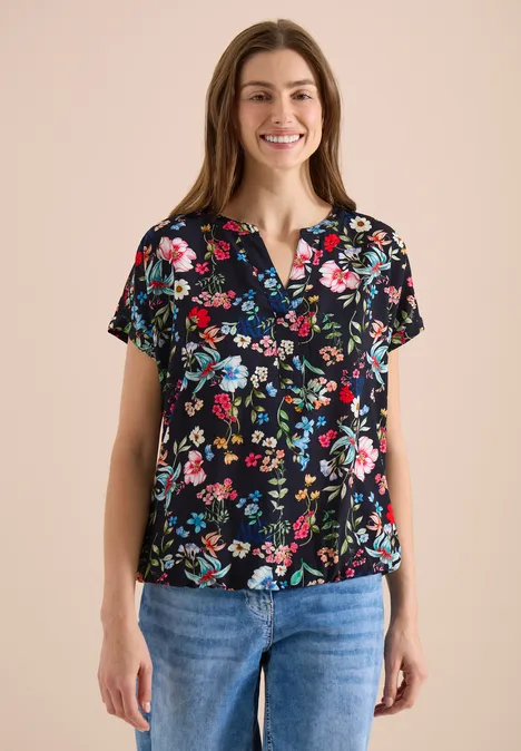 Frau in schwarzer Bluse mit buntem Blumenmuster und blauen Jeans, frontal stehend.