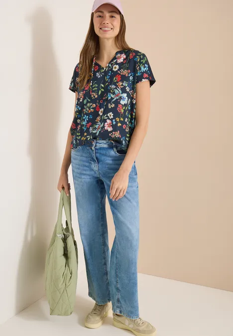 Frau in geblümter Bluse, blauen Jeans und Sneakern mit grüner Tasche vor hellem Hintergrund.